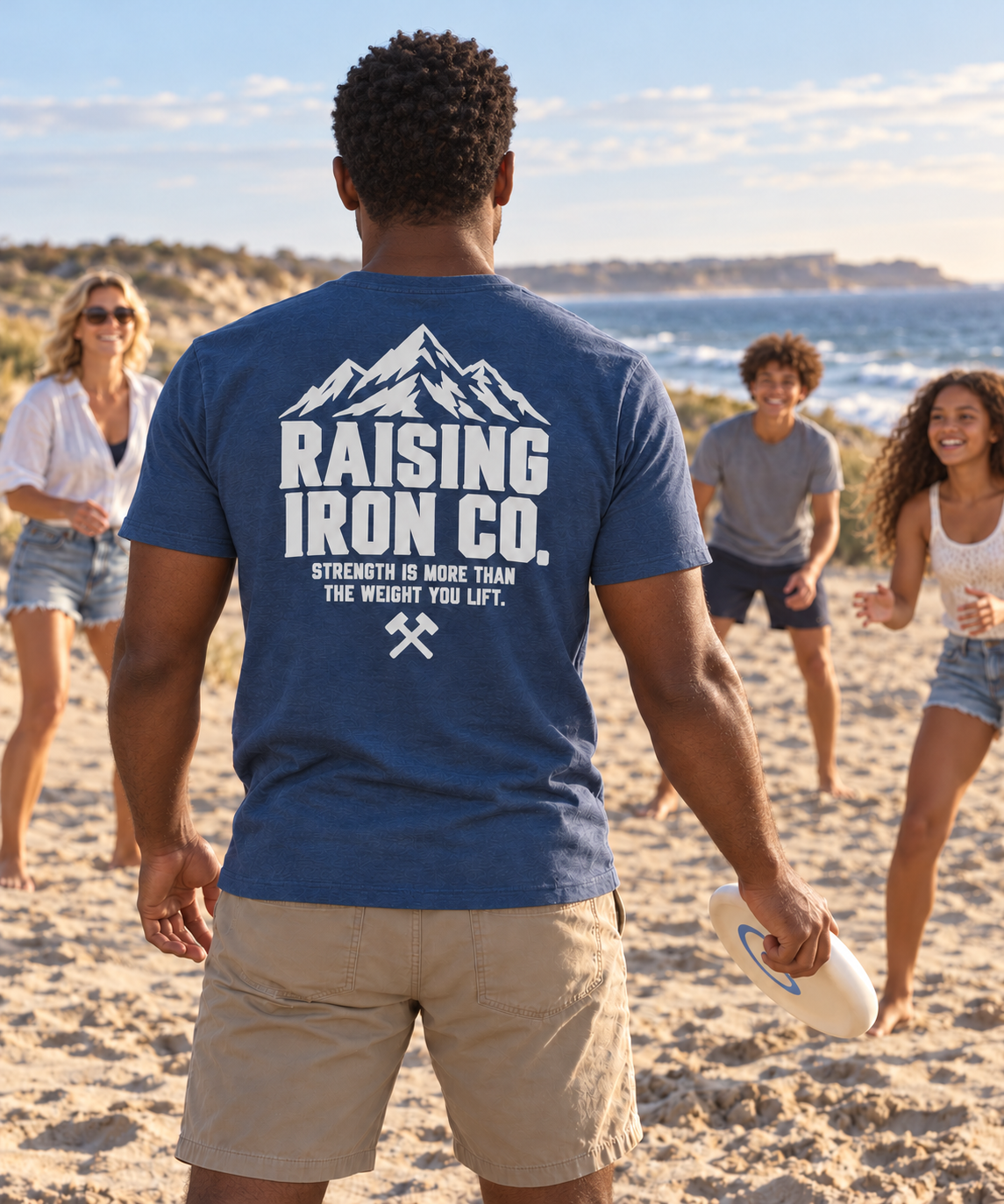 A man on a sandy beach stands with his back to the camera wearing a navy blue t-shirt with a large white graphic that reads 'RAISING IRON CO.' and 'STRENGTH IS MORE THAN THE WEIGHT YOU LIFT?' beneath a mountain illustration.
